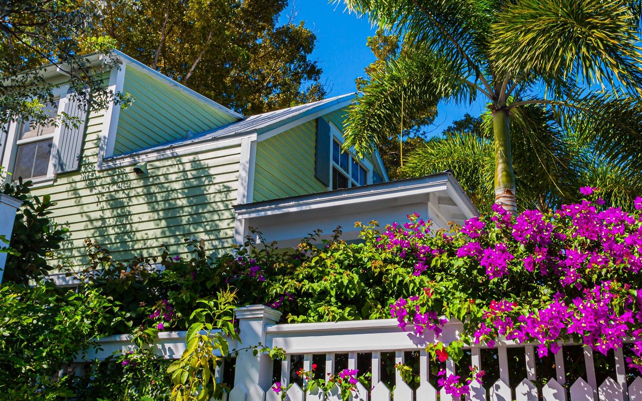 Light green two-story house with white trim, palms, and magenta bougainvillea behind a white fence.