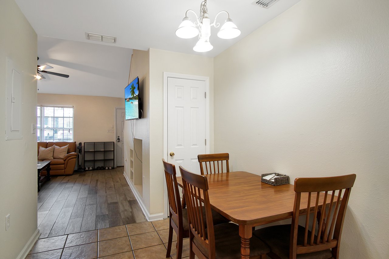 A cozy dining area with a wooden table and four chairs, adjacent to a living room with a tan sofa and wall-mounted TV, lit by a ceiling light.