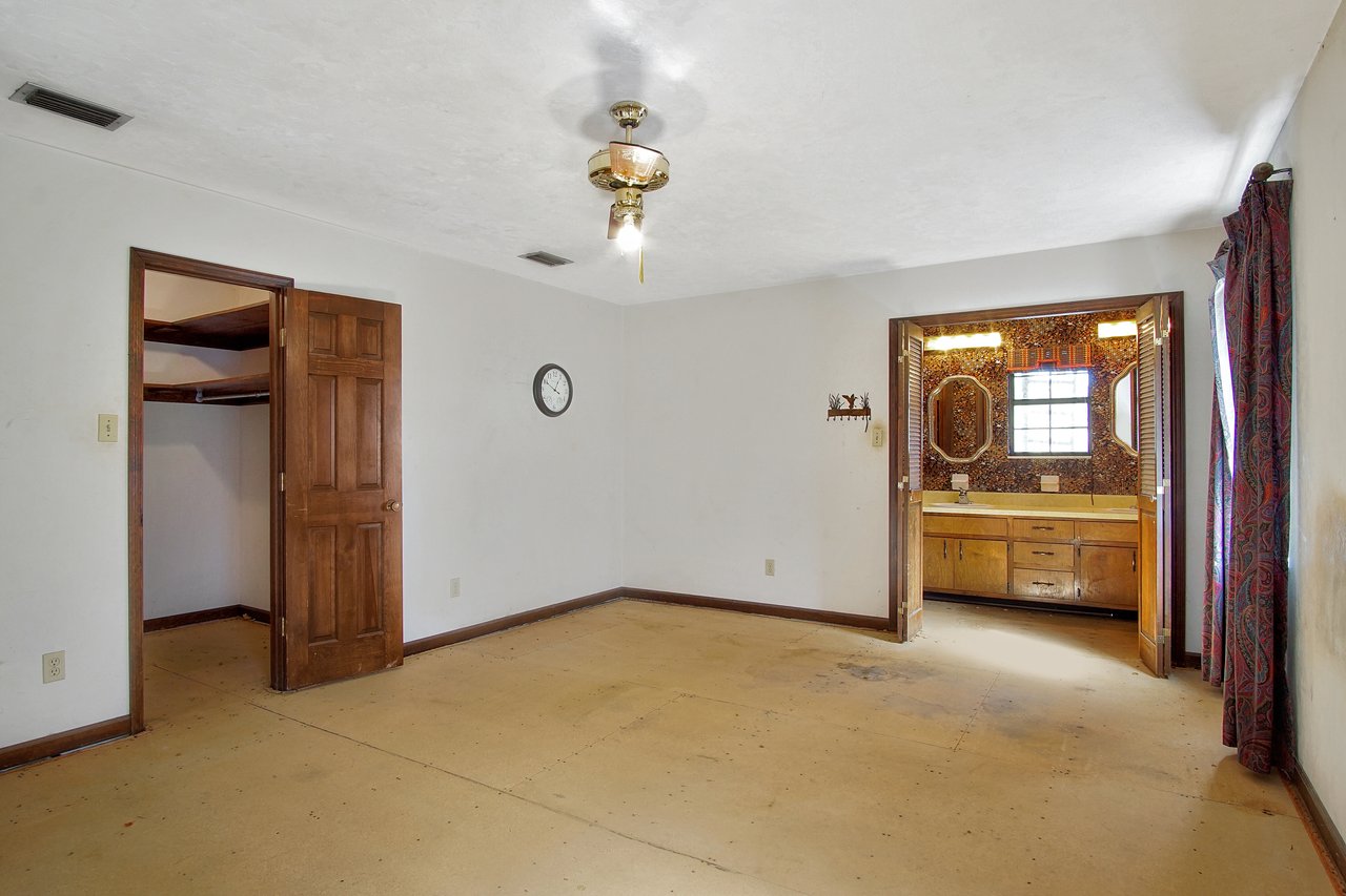 Empty room with beige carpet, wooden closet door, and wall clock. Right side opens to a bathroom with a wooden vanity, bright lights, and colorful curtains.