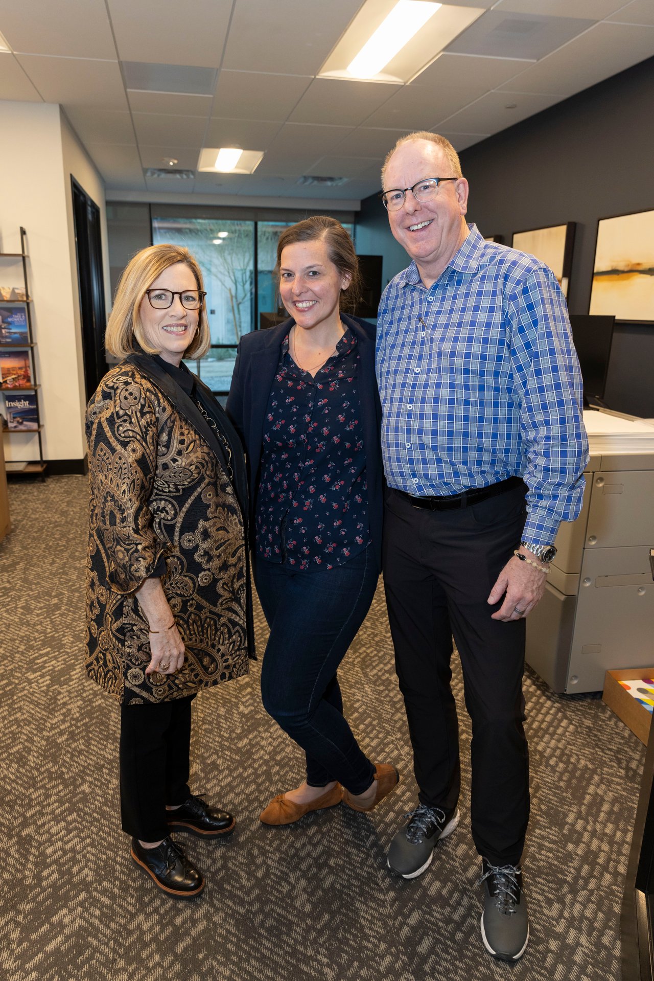 Three adults pose together in an office, including a man in a blue plaid shirt.