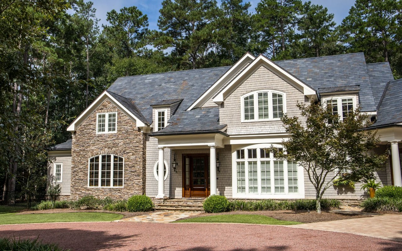 Large gray-shingled house with stone accents, a broad roof, and landscaped lawn among trees.