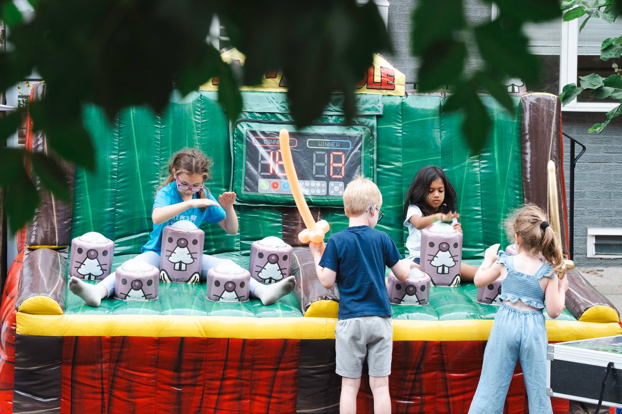 Children play a carnival game in a green inflatable booth at an outdoor block party.