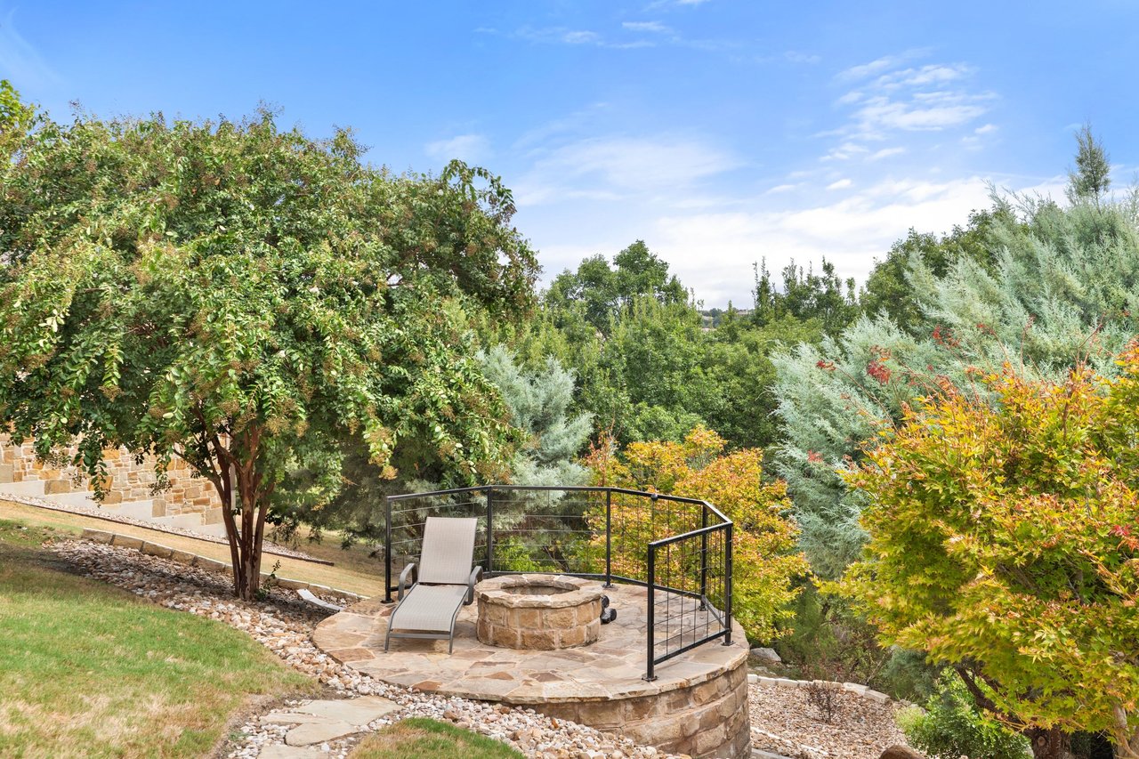 Landscaped stone patio with a circular fire pit, lounge chair, and surrounding trees.