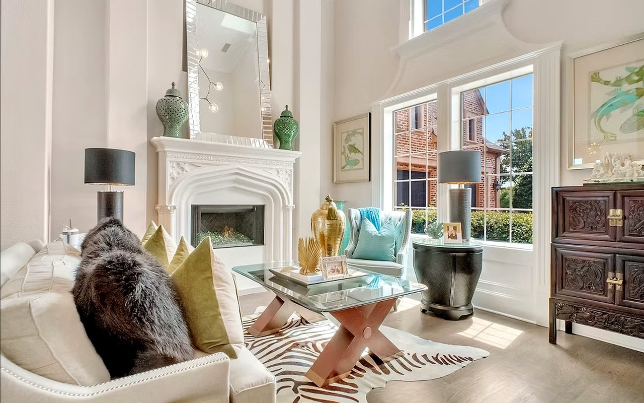 Living room with an ornate white fireplace, glass coffee table, and large sunlit windows.