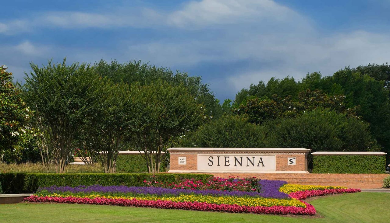 Sienna neighborhood entrance sign amid colorful flower beds and mature trees.