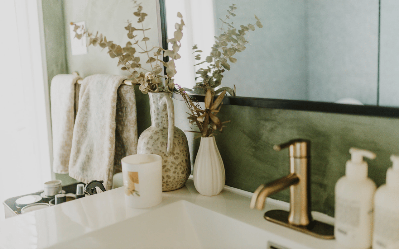 Bathroom vanity with a brass faucet, dark green backsplash, and vases of dried foliage.