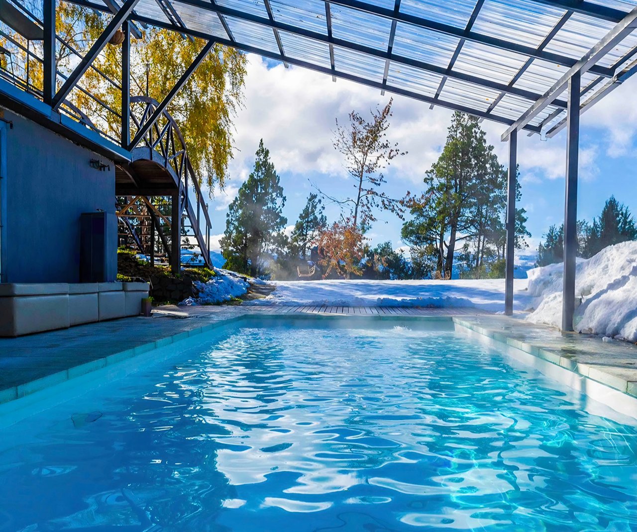 Outdoor pool beneath a glass canopy, with snow on the deck and trees beyond.