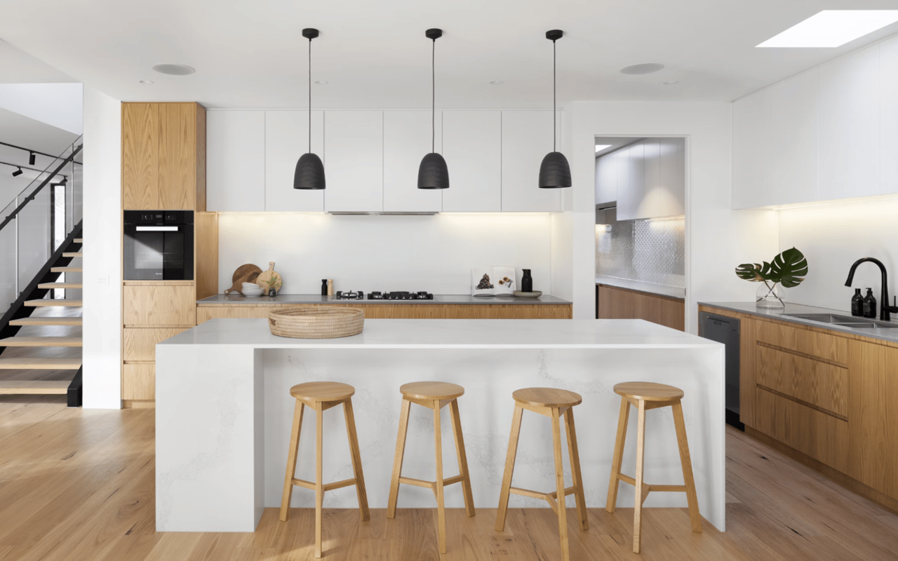  A spacious, modern kitchen features a large white island with a light-colored countertop and four wooden bar stools lined up in front of it. 