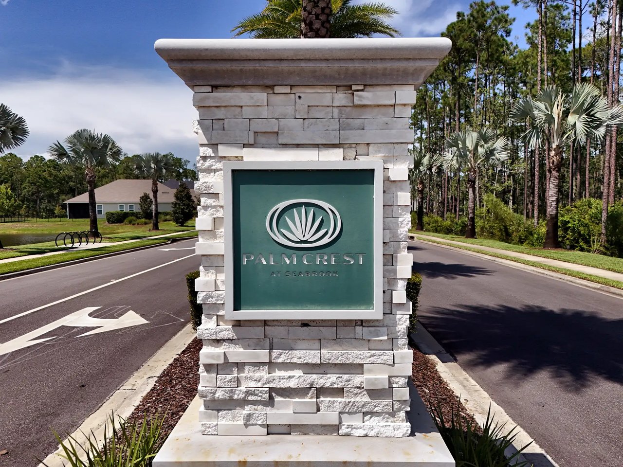 Palm Crest at Seabrook entrance monument with a teal palm-emblem sign on a white stacked-stone pillar, framed by palms and pines.