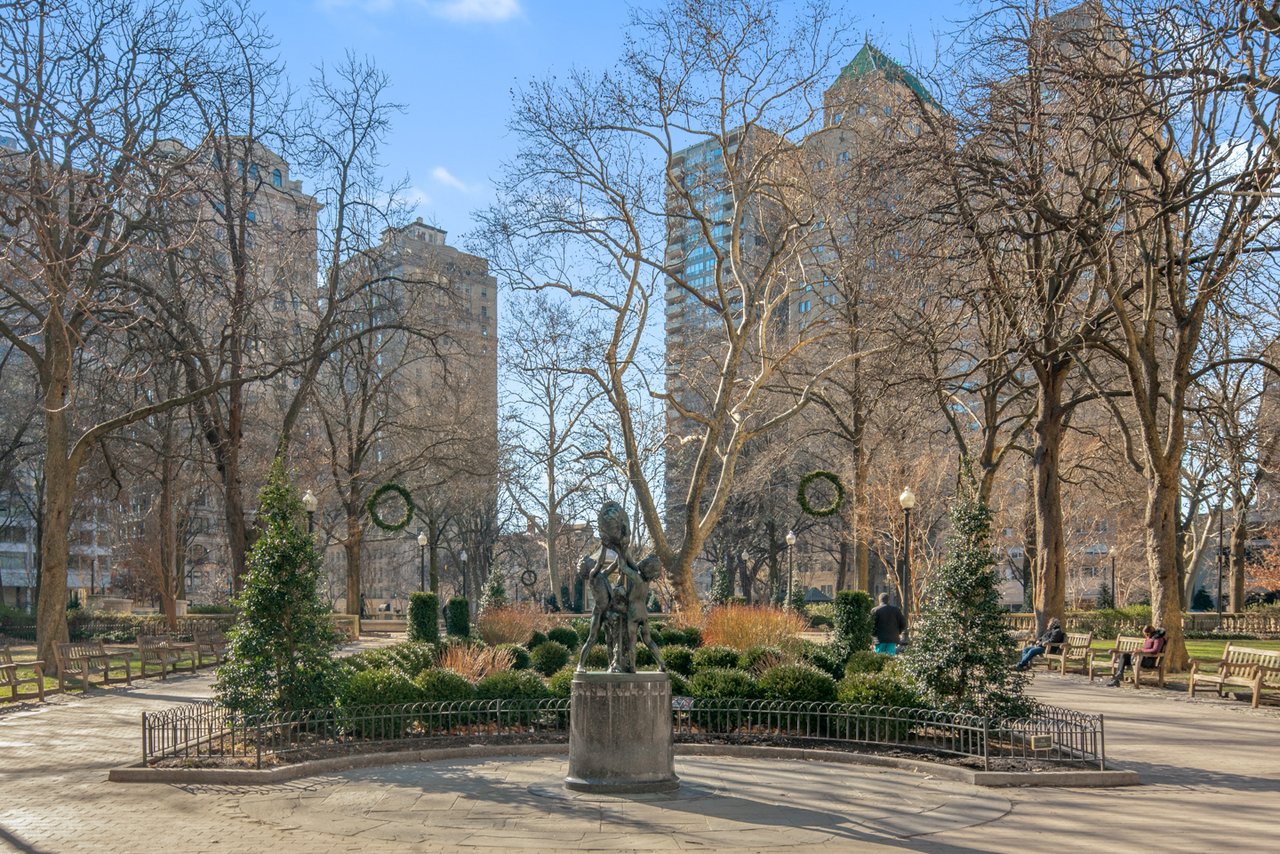 Urban park square with a bronze statue, benches, and tall buildings beyond leafless trees.