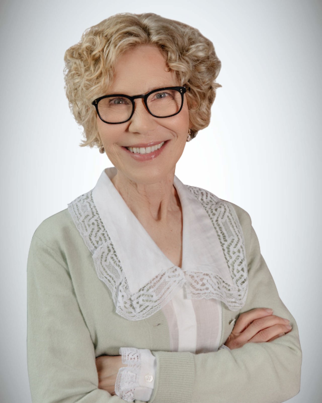 Debra Feldman smiles with her arms crossed, wearing black glasses and a pale green cardigan.