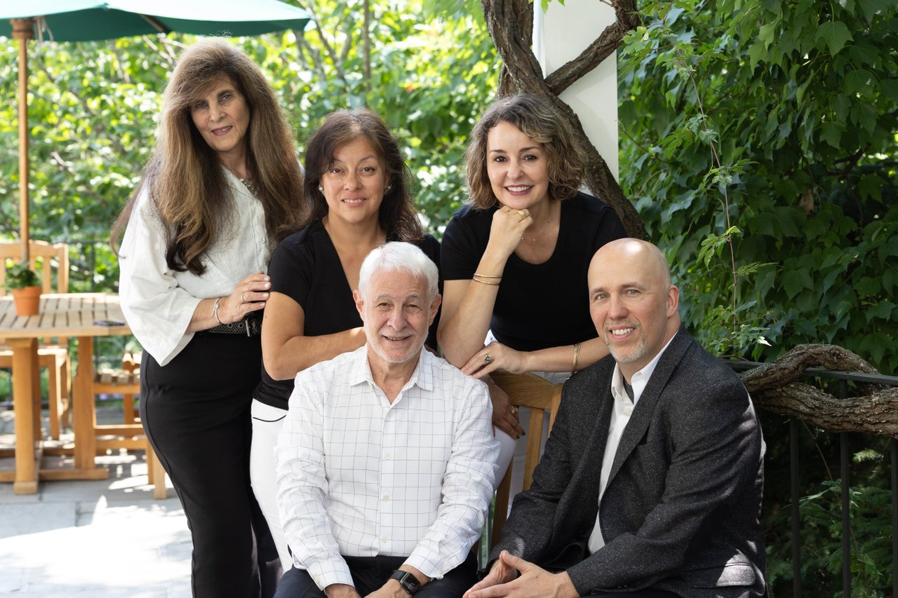 Five adults pose together on a leafy patio, three women standing behind two seated men.