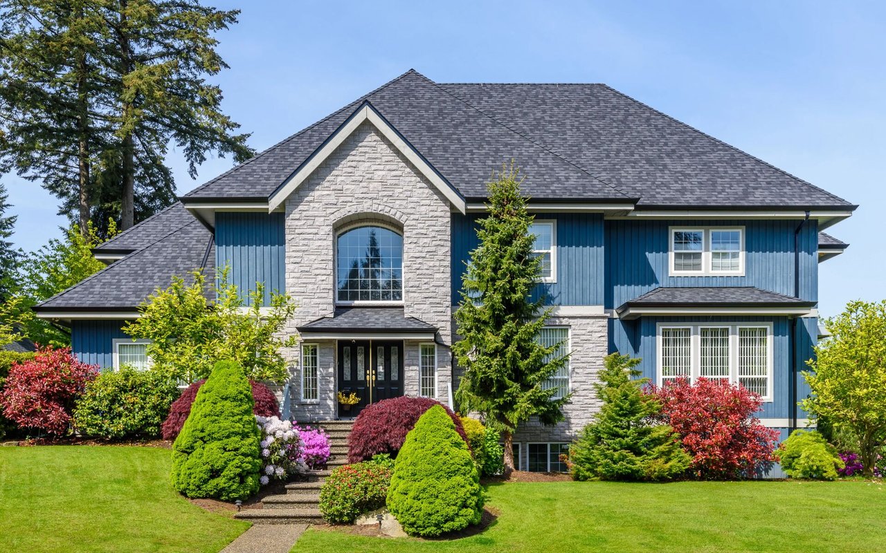 Two-story blue house with stone accents, a dark shingle roof, and colorful landscaped gardens.