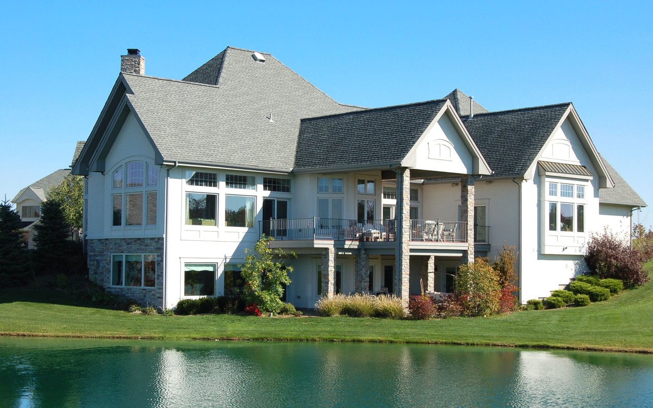 A large, elegant two-story house with light-colored siding and a grey shingled roof, situated beside a calm body of water