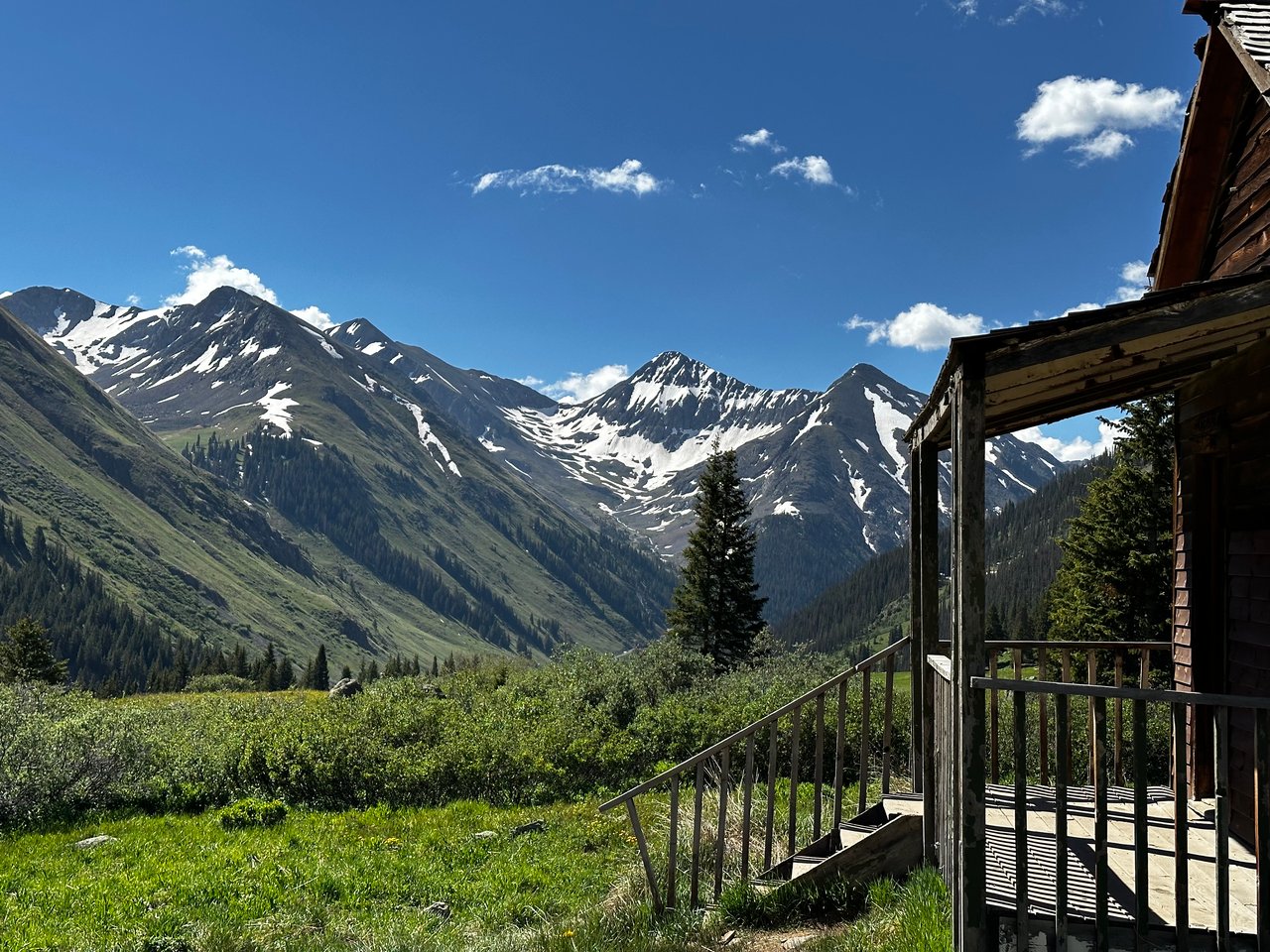 Rustic cabin porch overlooking snow-streaked mountains and a green alpine meadow.