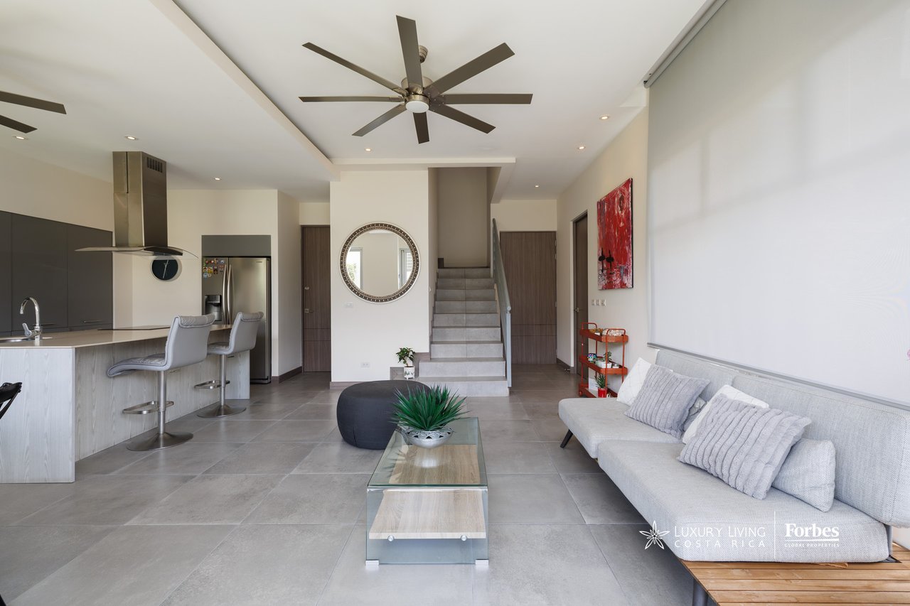 Open-plan townhouse living room with a gray sofa, kitchen breakfast bar, and staircase.