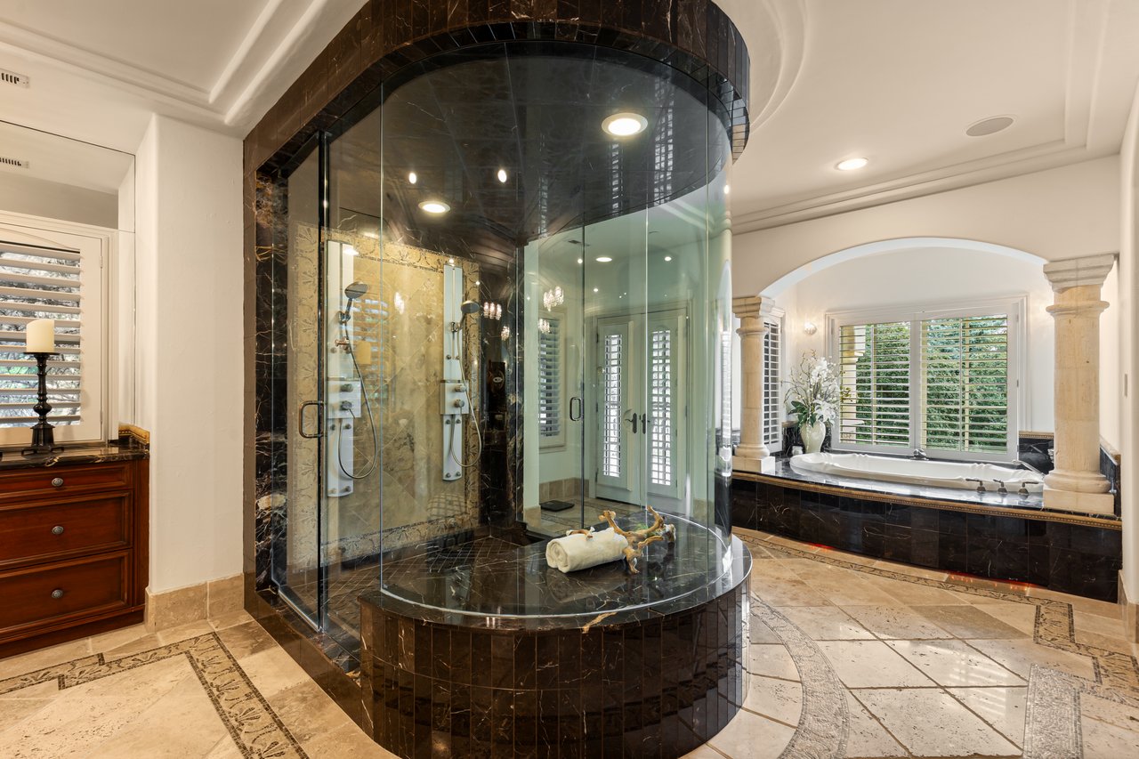 Luxury bathroom with a curved glass shower and a soaking tub beneath shuttered windows.