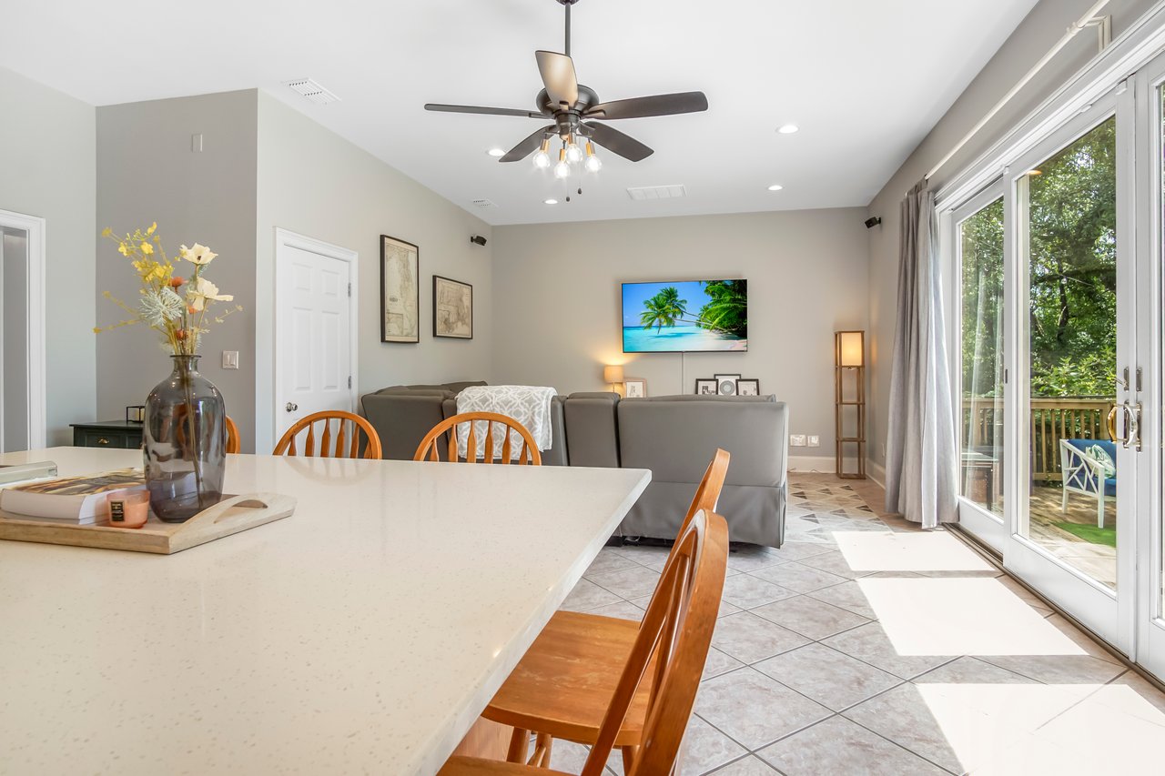 A bright, modern living room with a dining table and wooden chairs. A ceiling fan, large windows, and a wall-mounted TV create a cozy, inviting atmosphere.