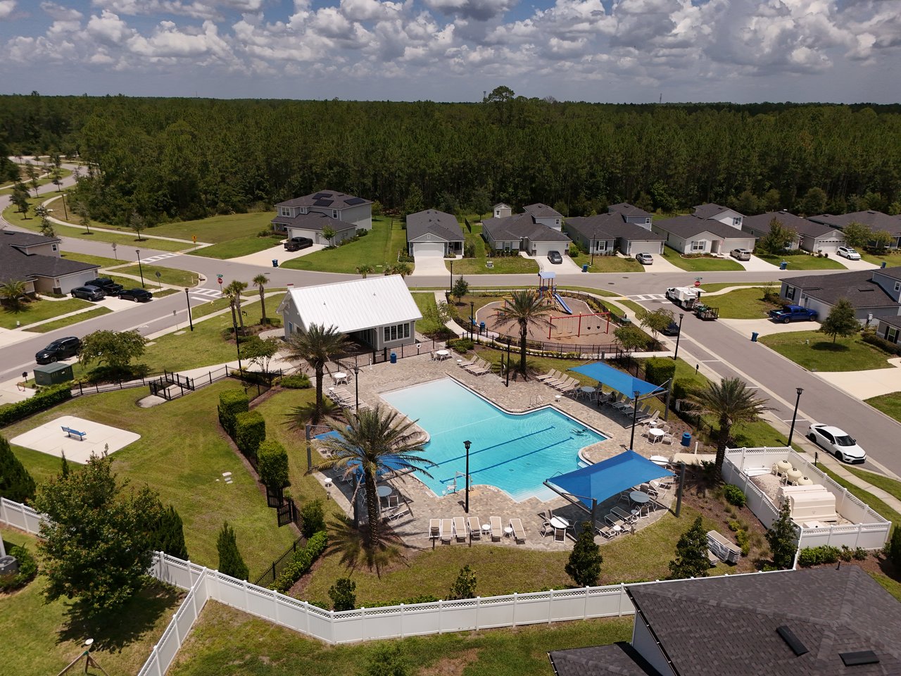 Aerial view of homes around a community pool, playground, and pavilion beside wooded land.