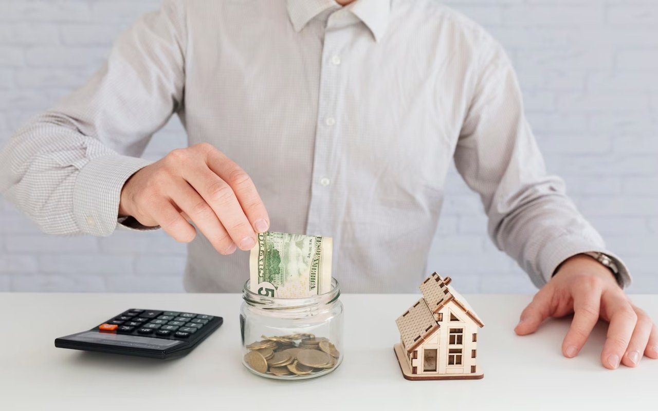 Person saves money in a jar beside a calculator and model house.