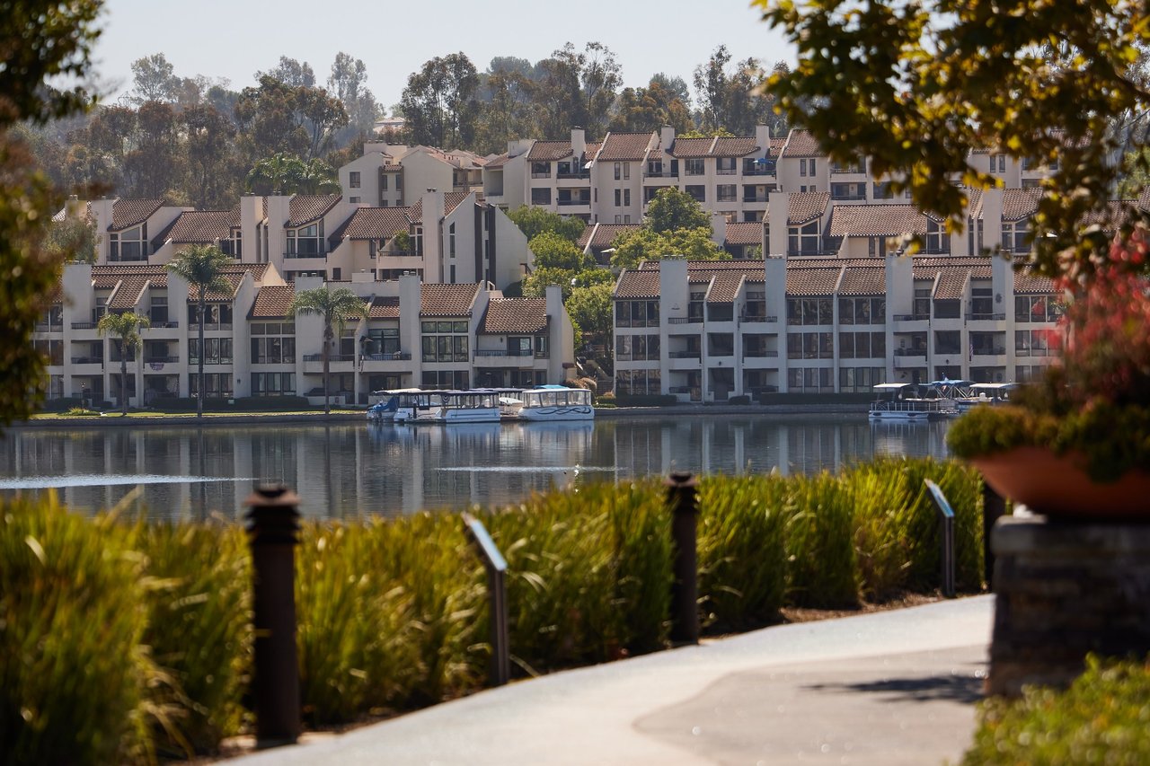 Curving paved walkway lined with tall ornamental grass and dark bollard lights beside a lake, with covered boats docked in front of tile-roofed waterfront buildings across the water
