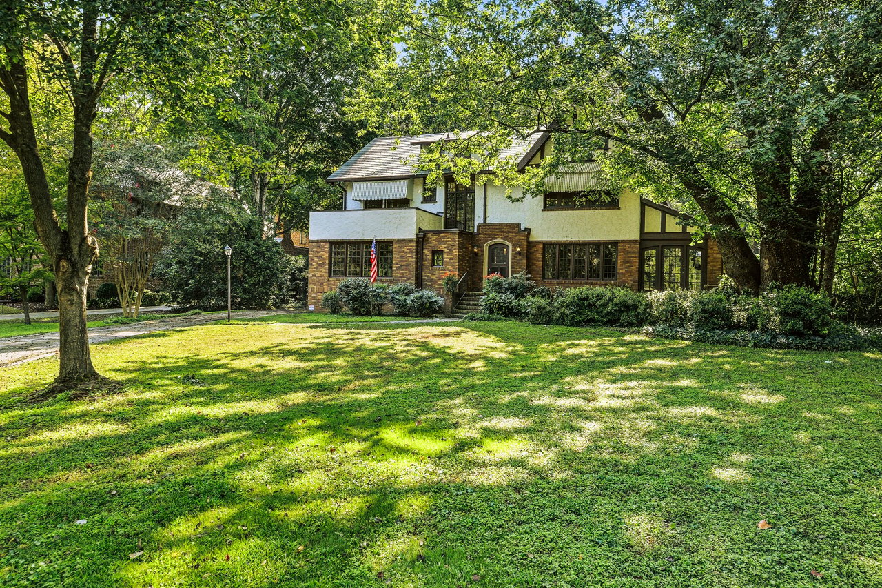 Two-story brick-and-white house set behind a sunlit lawn and mature trees.