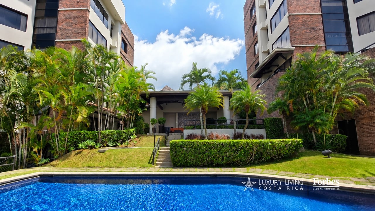 Luxury condominium courtyard with a blue pool, palm trees, and covered seating area.