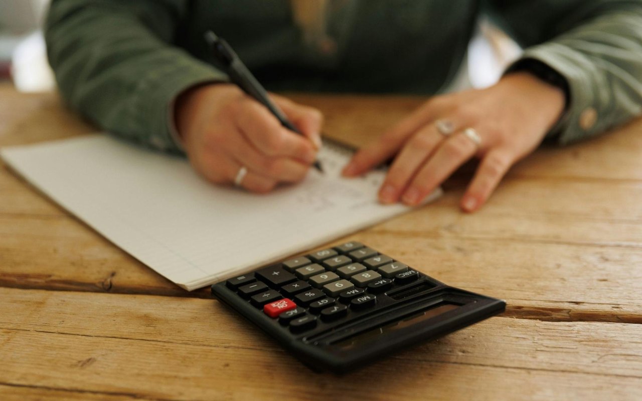  A person, seen from the chest up, is seated at a wooden table, writing on a piece of paper with a pen.