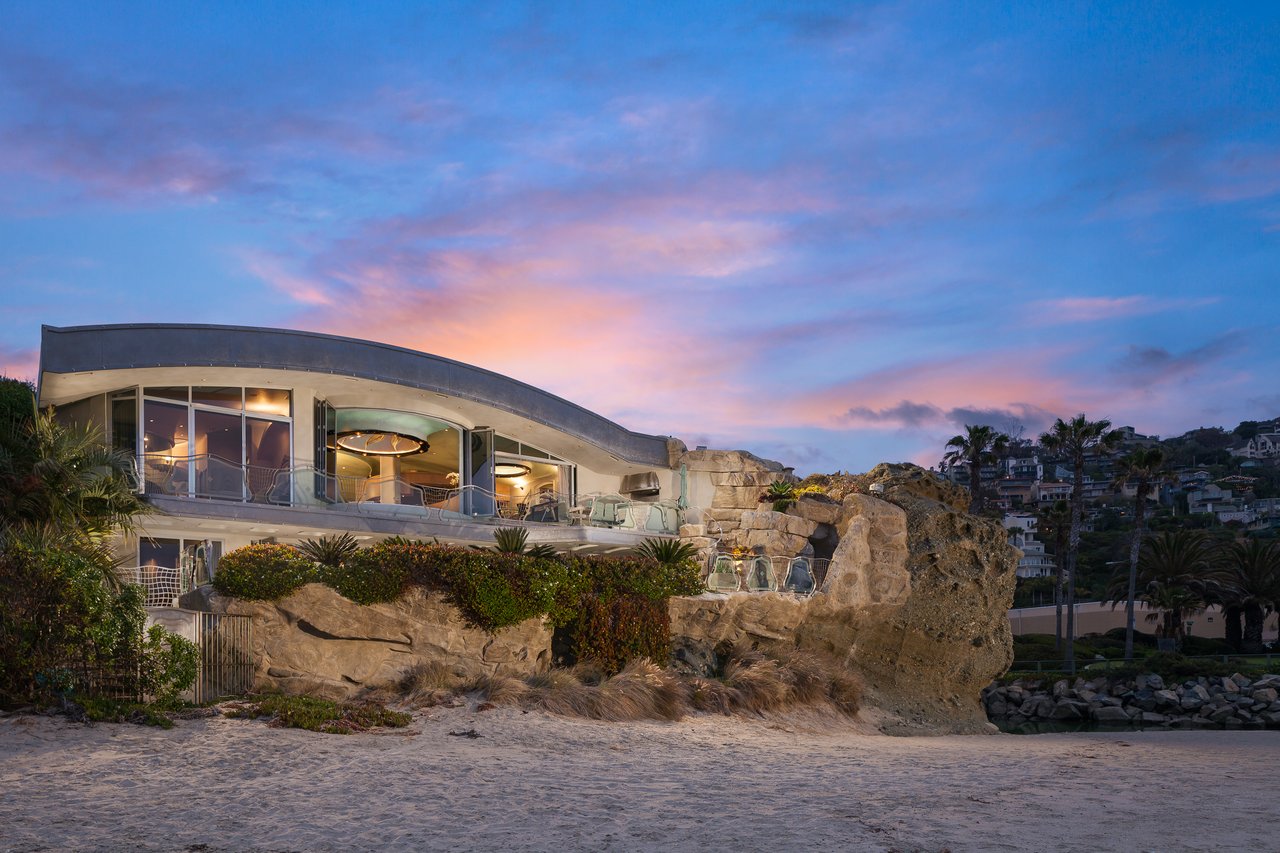 Modern beachfront house built into rocks, with a curved roof and glass walls at dusk.