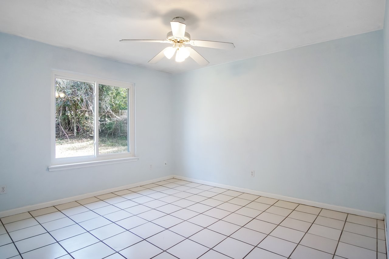 An empty room with light blue walls, white tile floor, and a large window showing greenery outside. A ceiling fan with a light fixture is centered. Bright and calm ambiance.