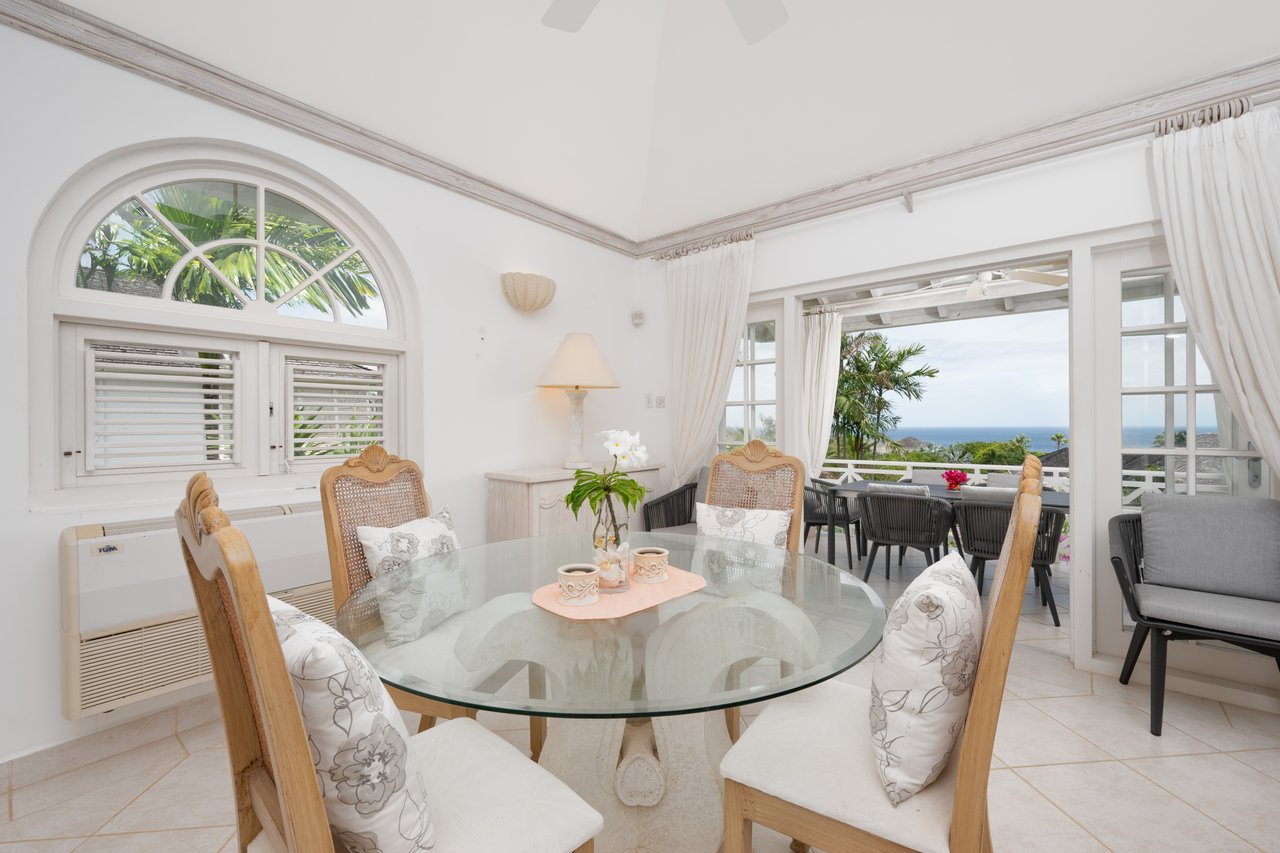 Dining room with a glass table, arched window, and open doors to a sea-view terrace.