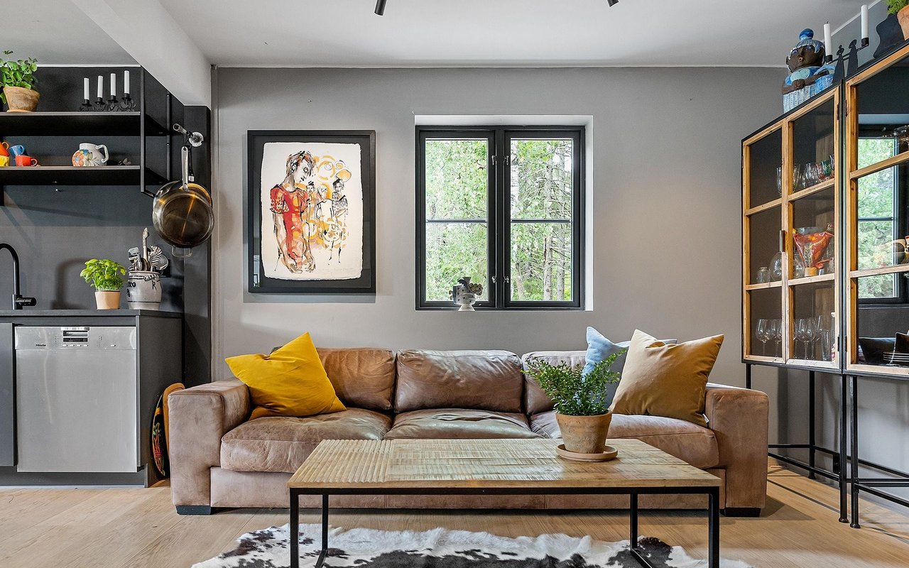 Open-plan living room with a brown leather sofa, colorful cushions, and a wooden coffee table beside a black kitchen.