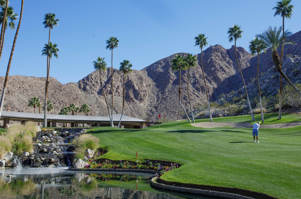 Golf course with palm trees, a pond, and rugged desert mountains.