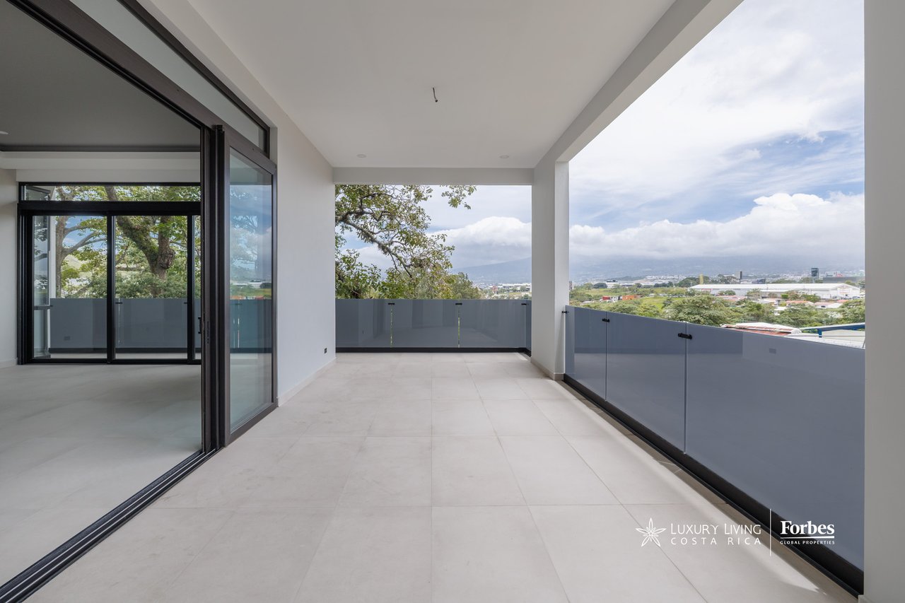 Covered apartment balcony with glass railing, tiled floor, and distant city and mountain views.