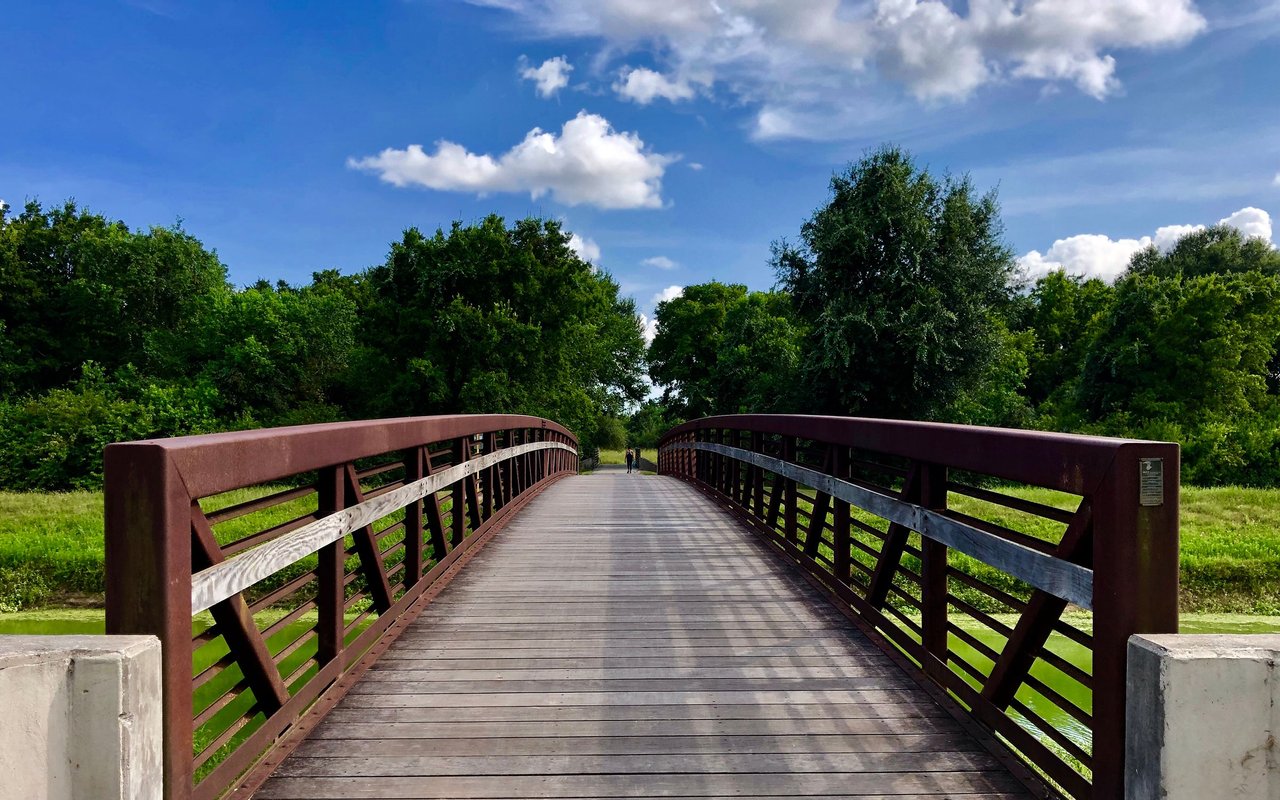 Wooden pedestrian bridge crossing a narrow waterway through a leafy park.