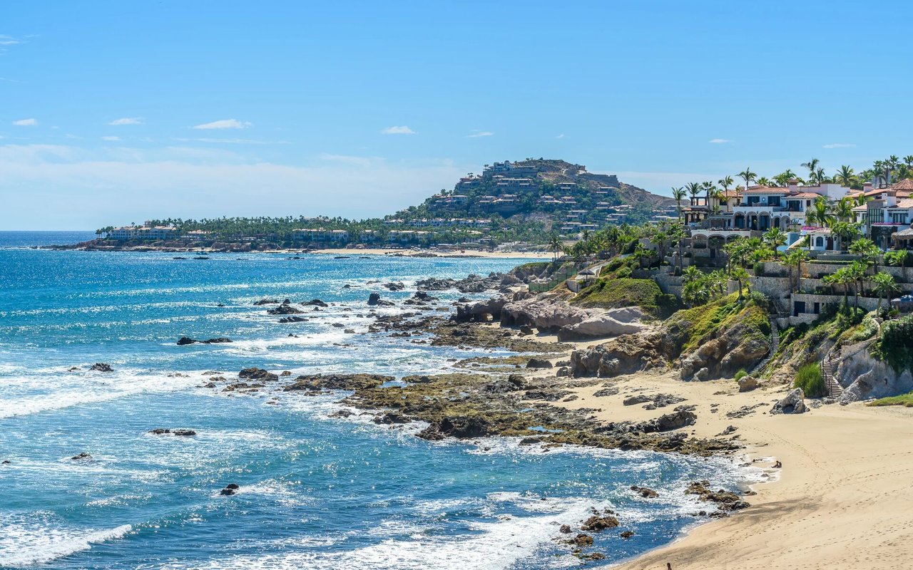 Rocky coastline with turquoise water, sandy beach, and palm-lined homes on a hillside.