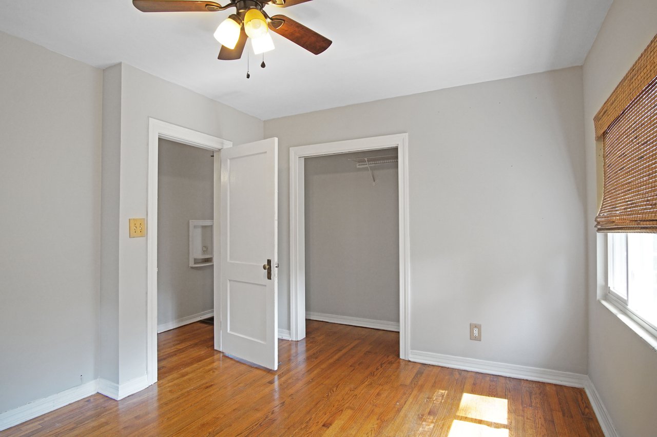 Empty room with wooden floor, white walls, open closet door, and ceiling fan. Sunlight streams through a window with bamboo blinds, creating a warm atmosphere.