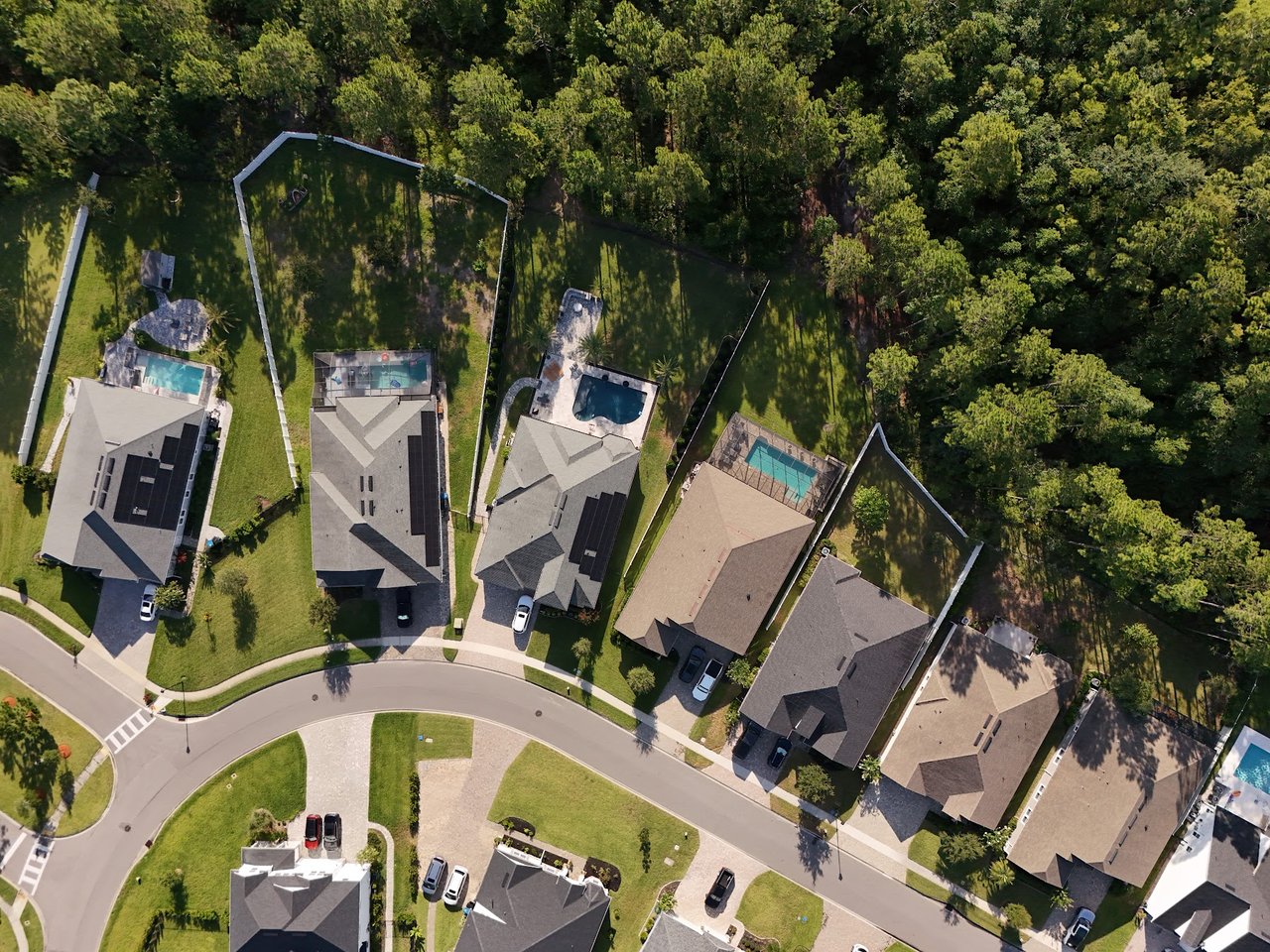 Aerial view of homes along a curved street, with backyard pools and mature trees.