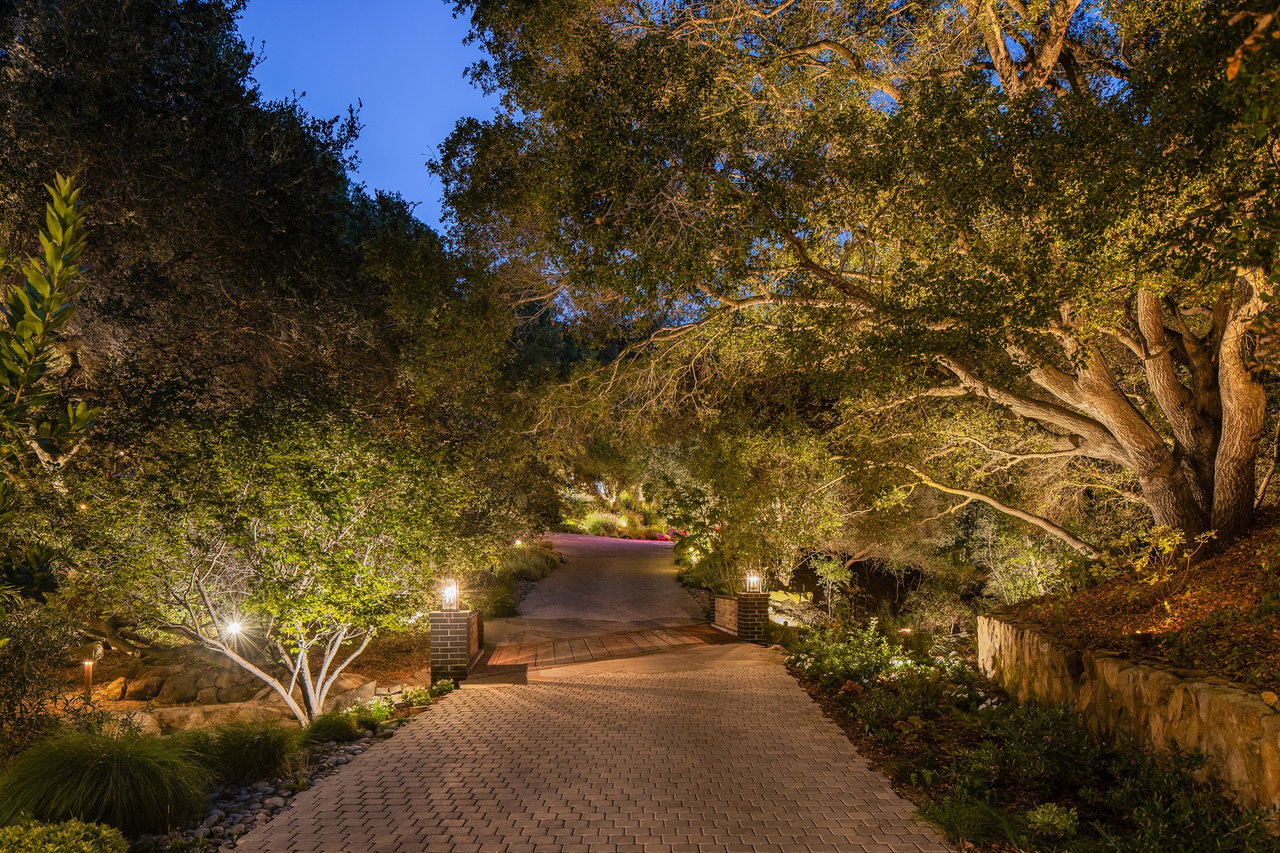 Brick driveway passes beneath mature trees, with garden lights illuminating the landscaped entrance at dusk.