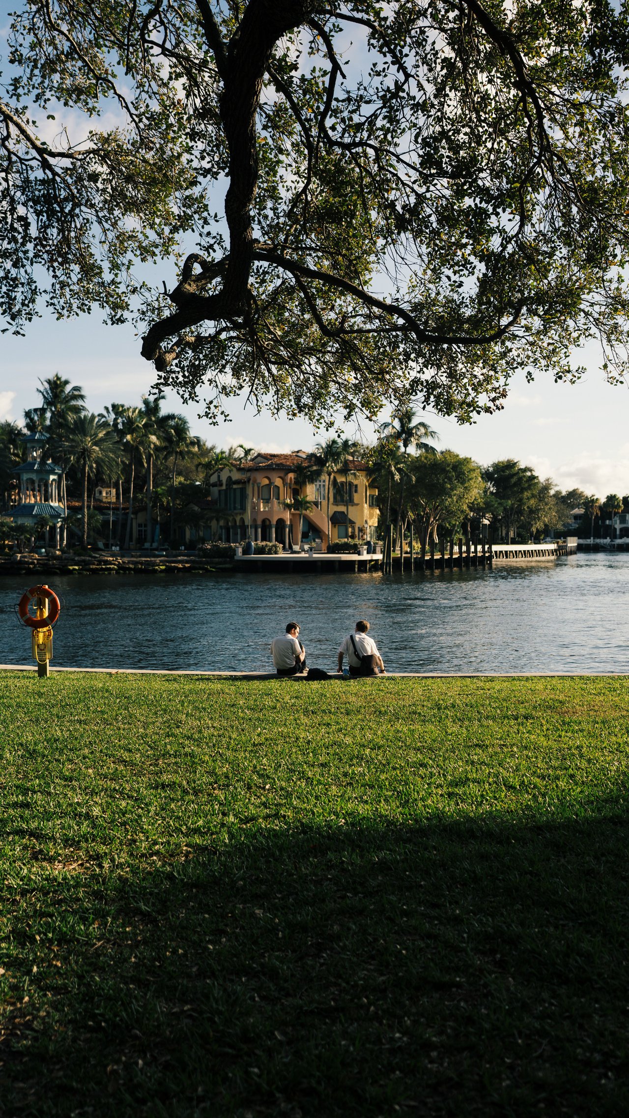Waterfront homes across a canal, framed by a large shade tree and two people sitting by the water.