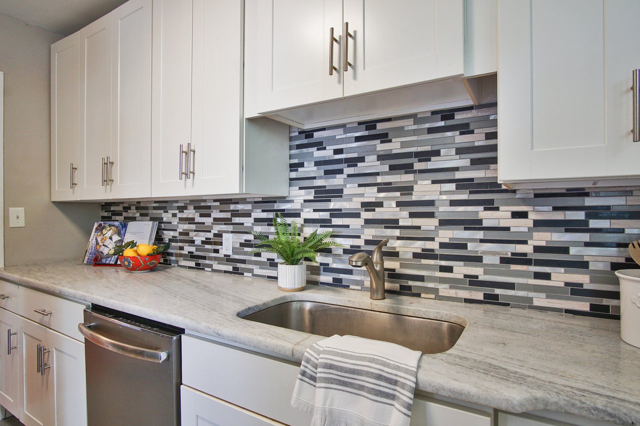 Modern kitchen with white cabinets, gray and black mosaic tile backsplash. Stainless steel sink, faucet, and dishwasher. Fruit bowl and plant add color.