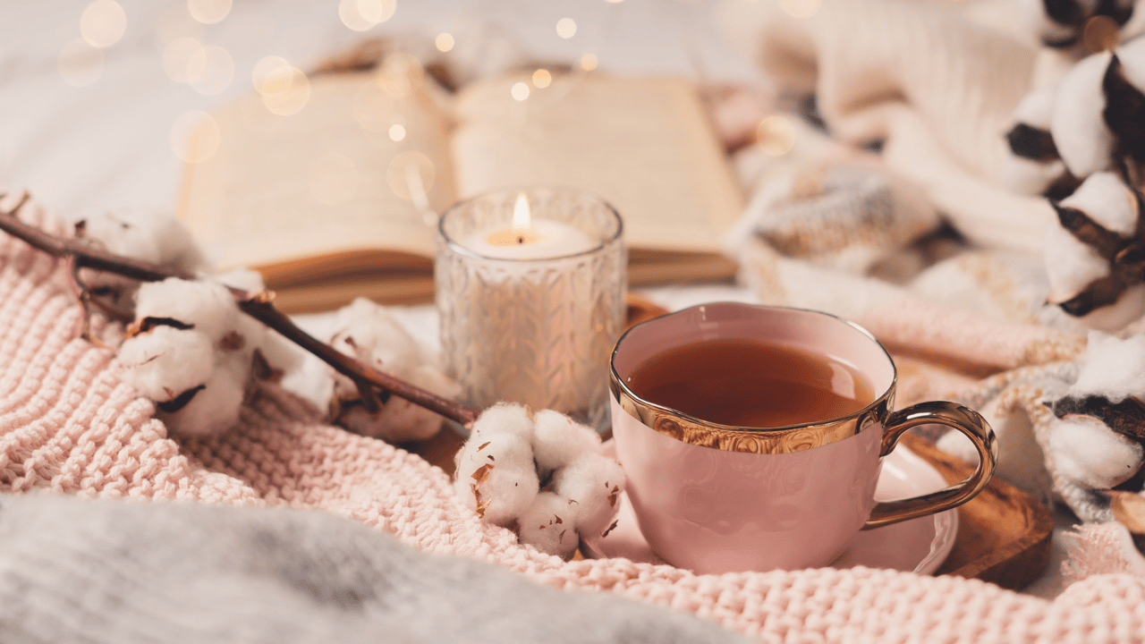 Cup of tea beside a candle and open book on a chunky knit blanket.