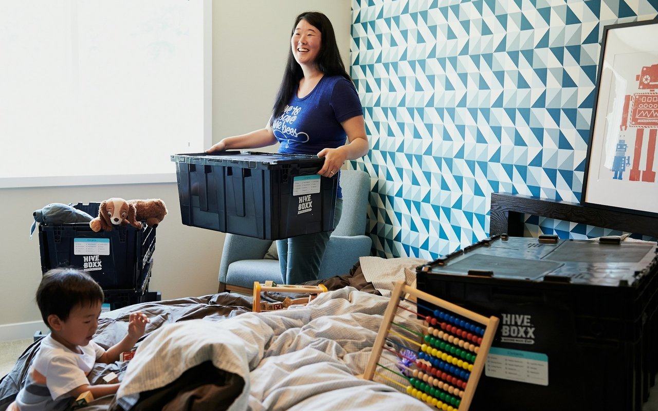 Woman carrying a moving bin as a child sits among packed belongings in a bedroom.