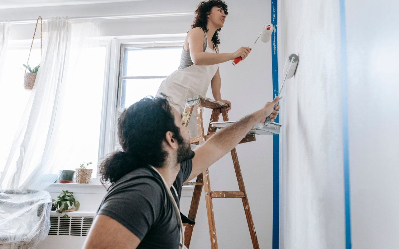 Two people paint a wall beside a wooden ladder in a bright room.