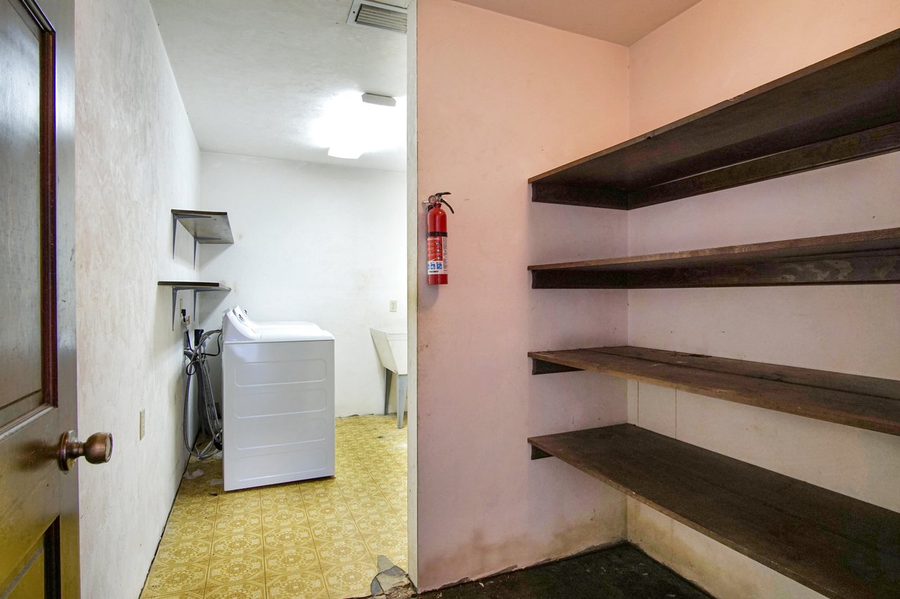 A small laundry room with a white washing machine on a patterned yellow floor. Empty dark wooden shelves on the right wall. A fire extinguisher is mounted near the ceiling under bright lighting.