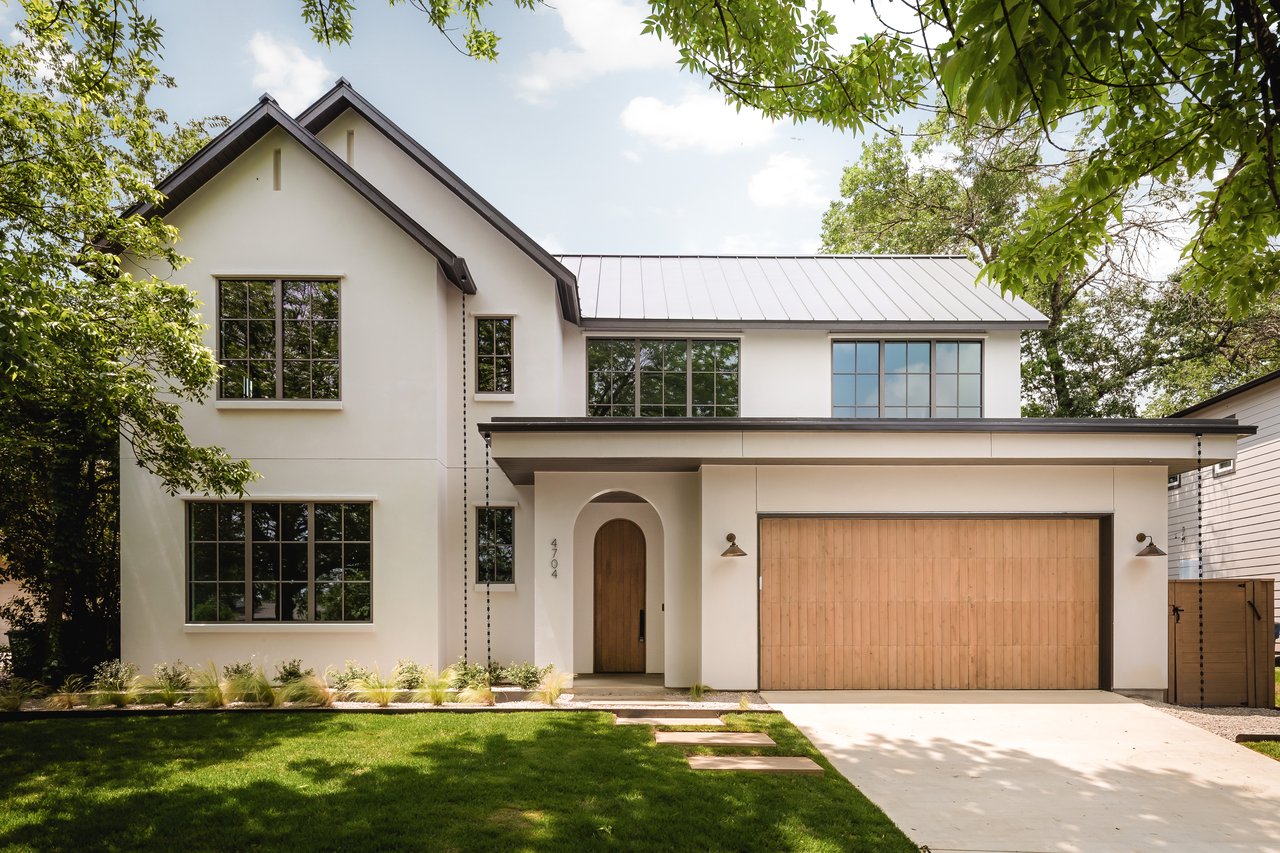 Modern white two-story house with an arched wood entry, wide wooden garage door, and leafy trees.