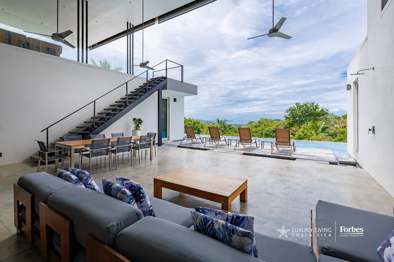 Modern outdoor living terrace with sofas, dining area, staircase, and pool overlooking tropical greenery.