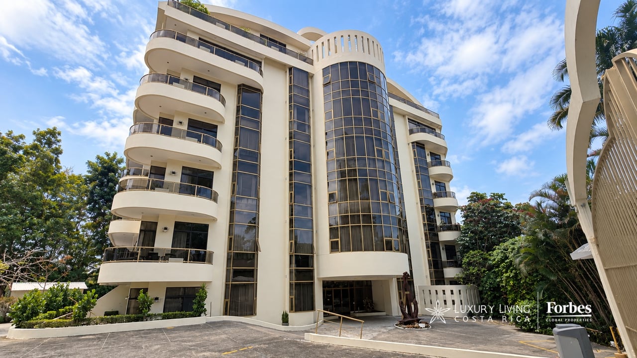 Cream-colored apartment building with curved balconies, tall glass windows, and tropical landscaping.