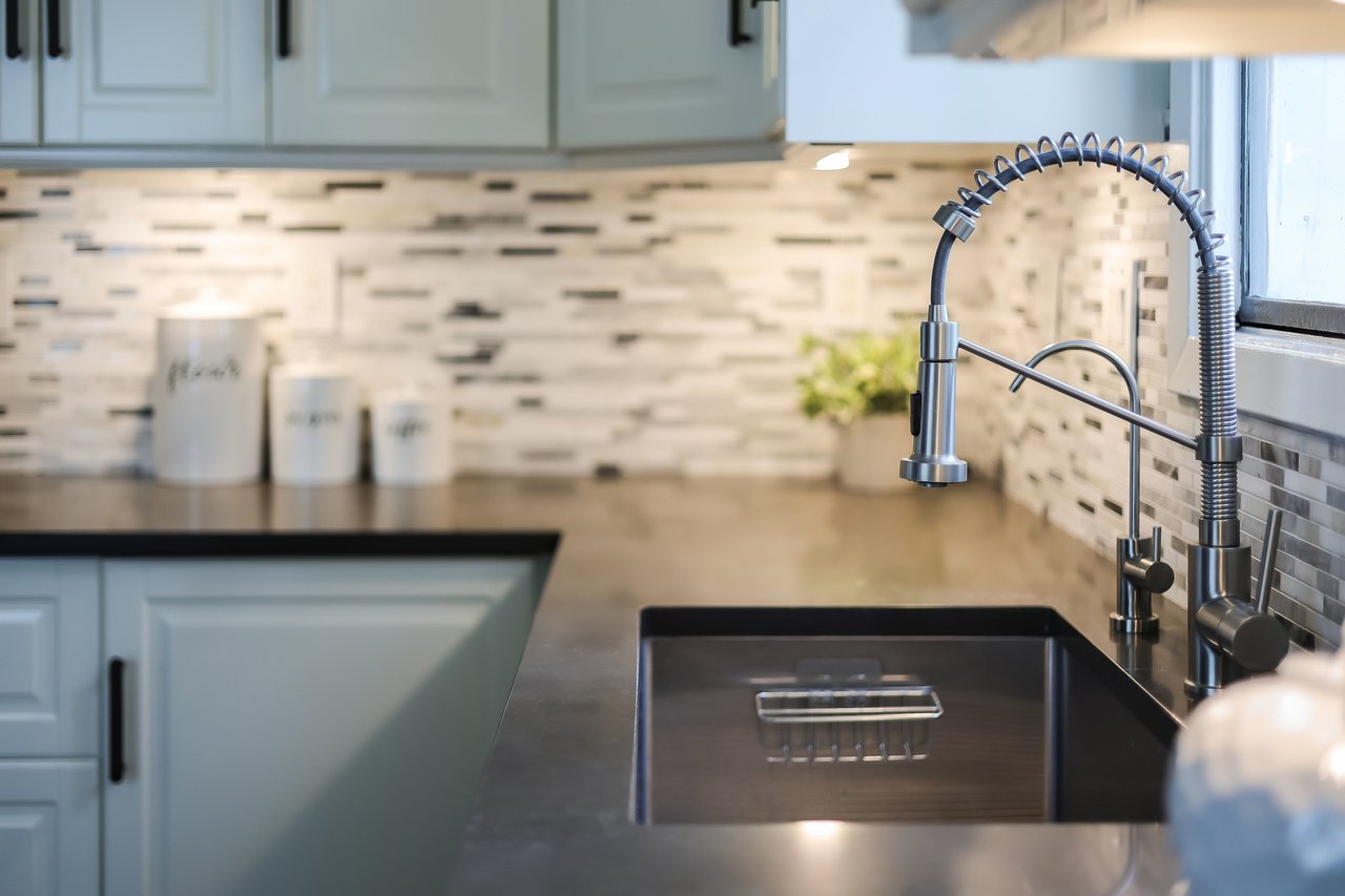 Kitchen sink with a spring-coil faucet, dark basin, gray cabinets, and mosaic tile backsplash.