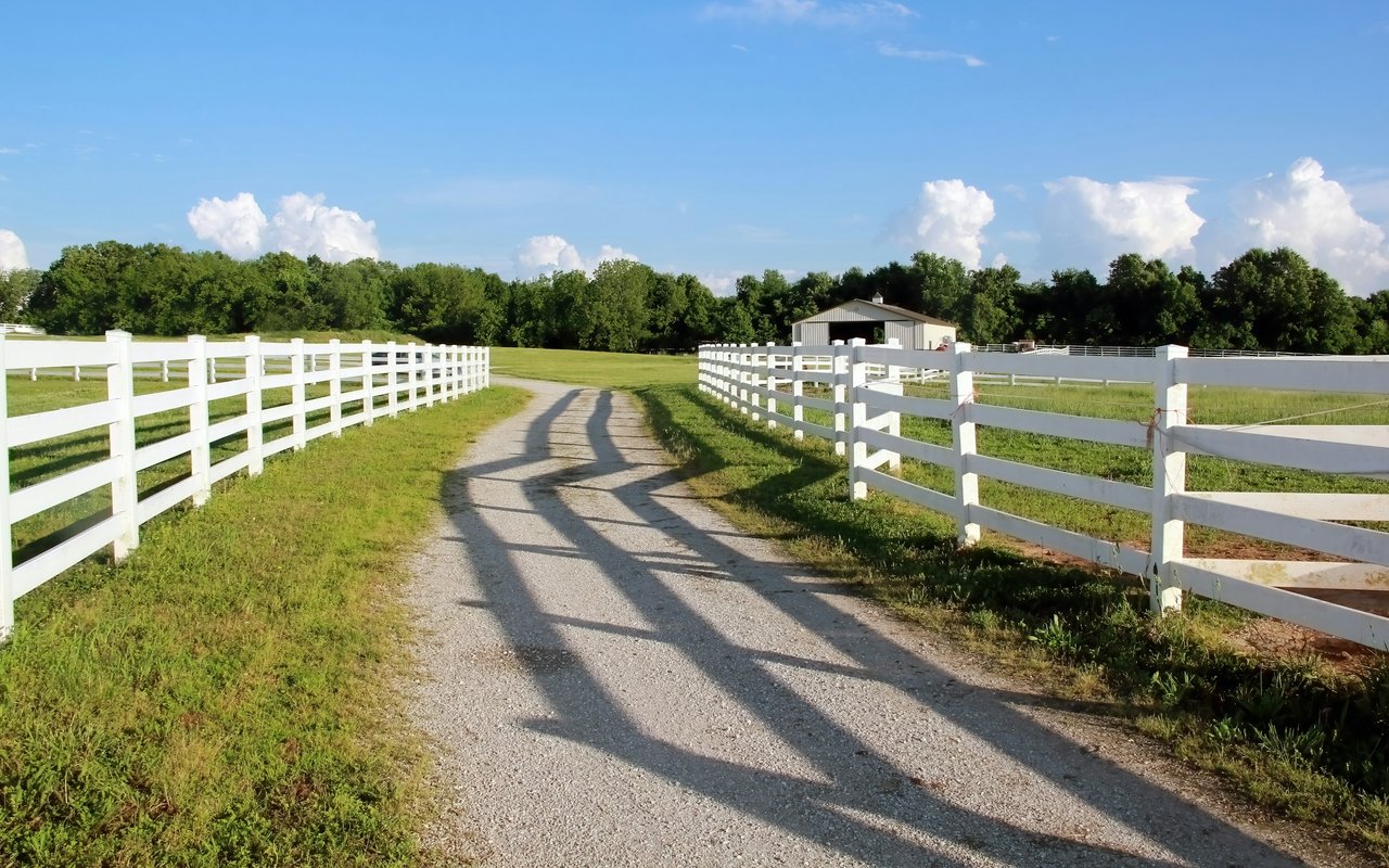 Gravel driveway between white fences leading toward a barn on a green rural property.